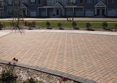 Townhouses with garages and balconies face a wide brick-paved courtyard with landscaped flower beds.