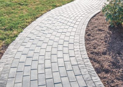 Curved stone walkway bordered by grass on one side and mulch with plants on the other.