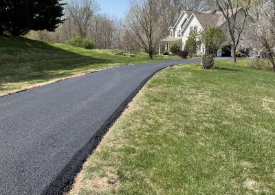 A newly paved driveway curves toward a white house surrounded by grass and trees under a clear blue sky.
