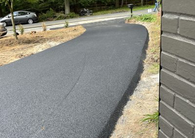 Freshly paved black asphalt driveway curves toward a road, bordered by trees and a brick wall.
