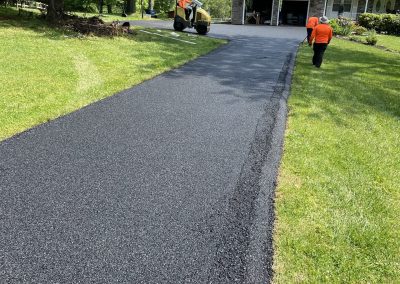 Freshly paved asphalt driveway with two workers near a house and green lawn on a sunny day.