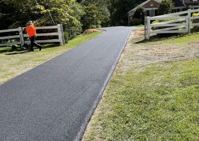 A person in an orange shirt works near a freshly paved driveway leading to a house with white fencing.