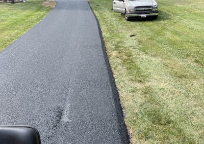 Freshly paved asphalt driveway with a car parked on the lawn and workers in the background near trees and a shed.