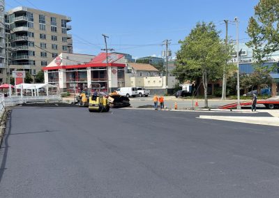 Freshly paved road with construction workers and equipment near a gas station and apartment buildings on a sunny day.