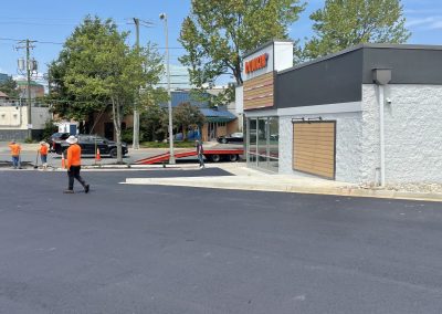Workers in orange shirts stand near a building and a sign on a freshly paved parking lot under a blue sky.