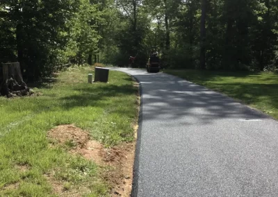 A newly paved road curves through a green, wooded area with sunlight filtering through the trees.