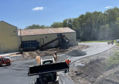 Construction vehicles pave a road near a yellow industrial building on a sunny day with trees in the background.