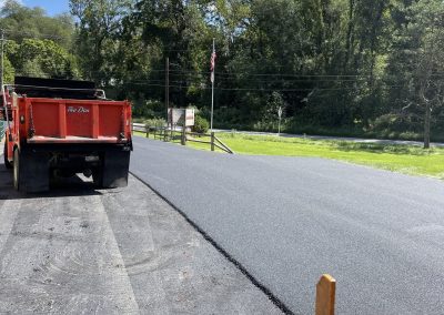 Fresh asphalt being laid on a road with construction vehicles and green trees in the background.