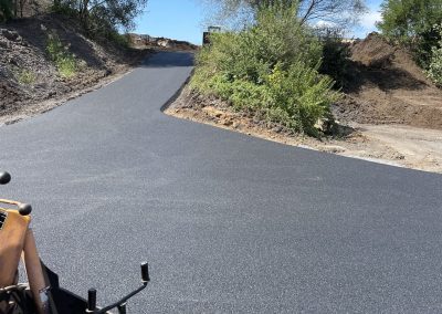 Freshly paved asphalt road curving uphill through a rural area with green trees and blue sky.