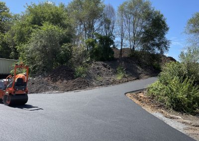Workers lay fresh asphalt on a curving road surrounded by trees under a clear blue sky.