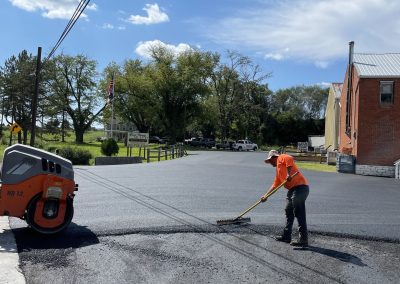 Worker in orange shirt spreading fresh asphalt in a parking lot on a sunny day with trees and buildings nearby.