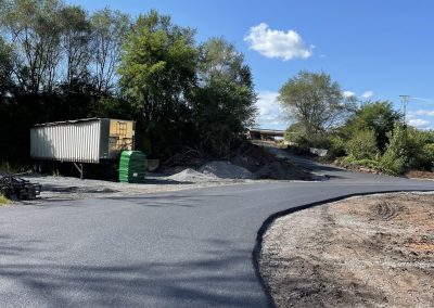 A freshly paved road curves past a shipping container and piles of gravel on a sunny day with blue sky.