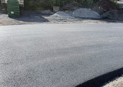 Freshly paved asphalt road with construction materials, gravel, and trees in the background on a sunny day.