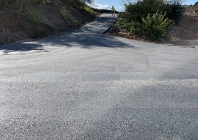Freshly paved road with a worker standing at the top of a hill, surrounded by trees under a blue sky.