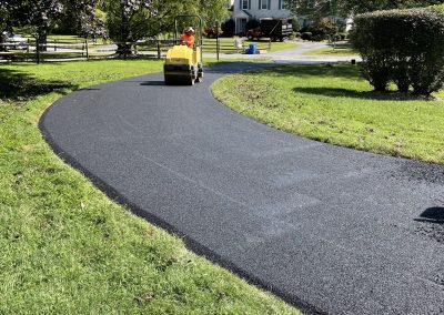 A worker uses a yellow roller to pave a curved asphalt driveway on a sunny day.