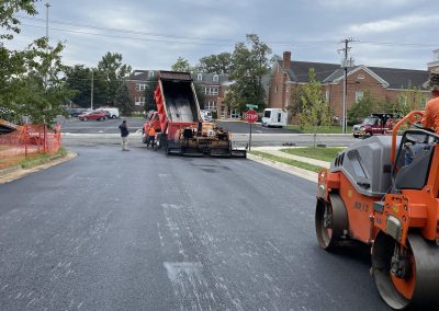 Fresh asphalt being laid on a road with construction vehicles and a worker nearby under a cloudy sky.
