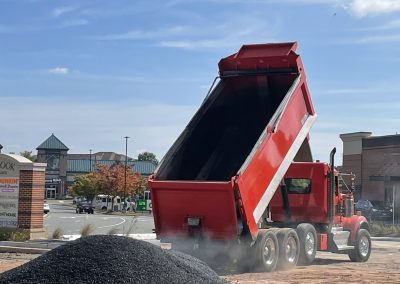 A red dump truck unloading gravel onto a construction site near shops on a sunny day.