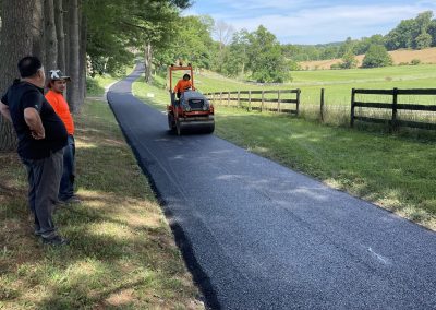 Workers observe as fresh asphalt is rolled onto a rural path lined with trees and wooden fences on a sunny day.