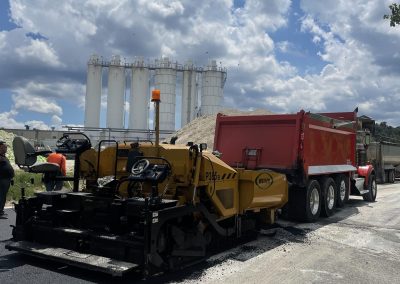A road paving machine and a red dump truck work on fresh asphalt under a partly cloudy sky.