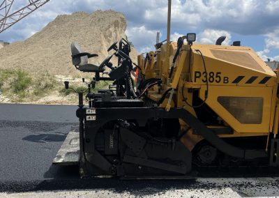 Yellow asphalt paver machine laying new blacktop on a road, with sand piles and cloudy sky in the background.