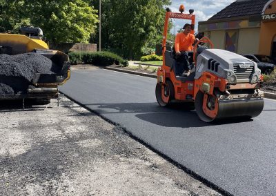 A worker operates a road roller on freshly paved asphalt near a commercial building on a sunny day.