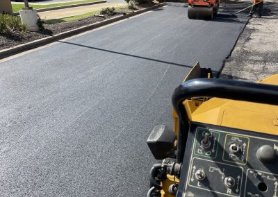 Workers paving a parking lot with fresh asphalt; machinery and control panel visible in the foreground.