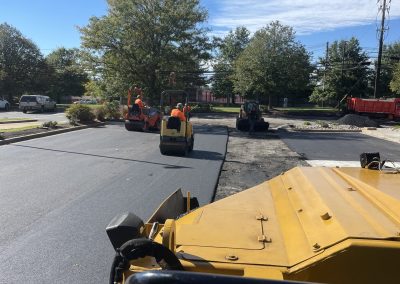 Construction workers use heavy machinery to pave a new road under a clear blue sky with trees in the background.