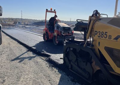 A road roller and paver machine lay fresh asphalt on a sunny day at a road construction site.