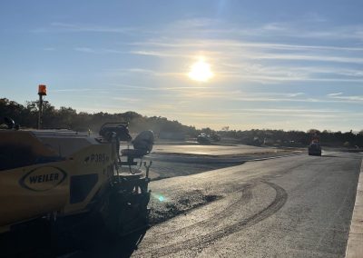 Sunset over a freshly paved road, with construction equipment and trees in the background.