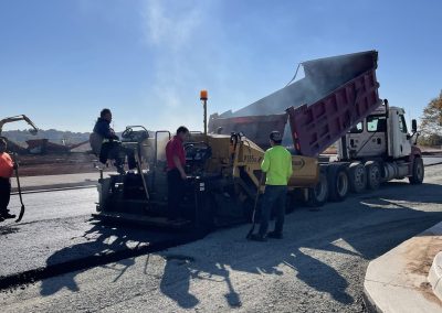 Workers operate heavy machinery to lay asphalt on a road under bright sunlight, with a truck dumping materials.