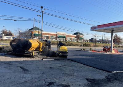 Two workers pave asphalt near an Exxon gas station with a yellow road roller on a sunny day.