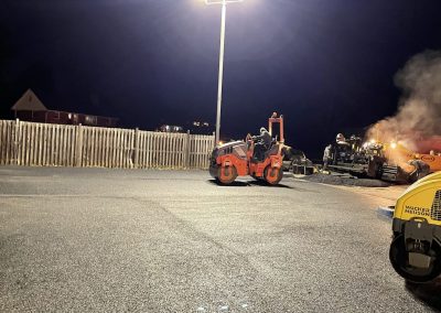 A parking lot being paved at night under bright floodlights, with workers and machinery visible.