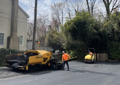 Two workers repaving a street with heavy machinery near trees and a house on a clear day.
