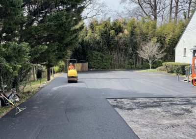 A worker drives a roller on freshly paved asphalt in a driveway surrounded by trees and a house.