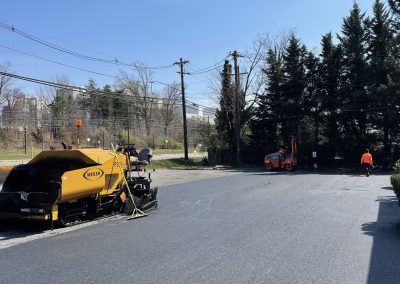 A fresh asphalt road is being paved with construction equipment and two workers under a clear blue sky.
