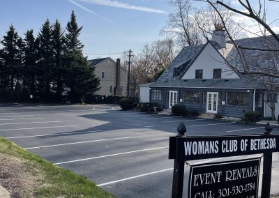 A white building with a sign for the Womans Club of Bethesda and an empty parking lot on a sunny day.