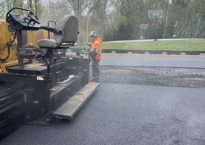 A worker in a safety vest stands next to paving equipment on a newly asphalted outdoor court.
