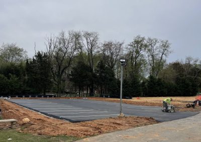 Workers pave a new parking lot near trees under a cloudy sky; construction equipment is visible.