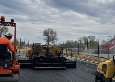 Workers paving a new road with heavy machinery under a cloudy sky, surrounded by fencing and trees.