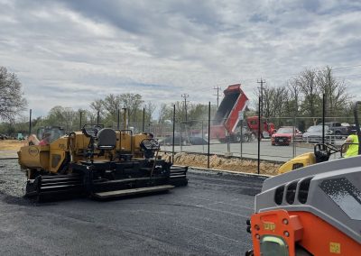 Construction vehicles pave a road on a cloudy day, with a red dump truck in the background.