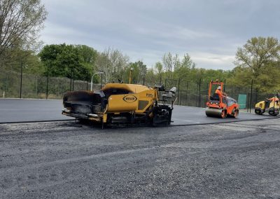 Yellow paving machines lay asphalt on a road surrounded by trees under a cloudy sky.
