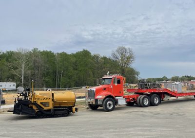 A yellow paving machine and a red semi-truck with a flatbed trailer parked at a construction site.