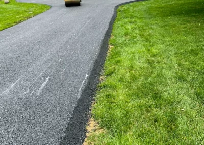 A person drives a steamroller, paving a fresh asphalt driveway next to a green lawn.