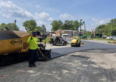 Workers in safety vests pave a road with heavy machinery under a partly cloudy sky.