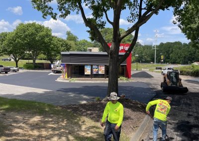 Workers pave a parking lot near a Wendy's restaurant on a sunny day, with trees providing some shade.