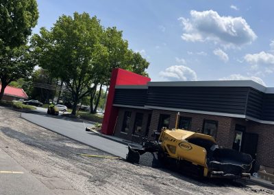 Yellow paving machine resurfacing a parking lot outside a building with red and gray accents under a blue sky.