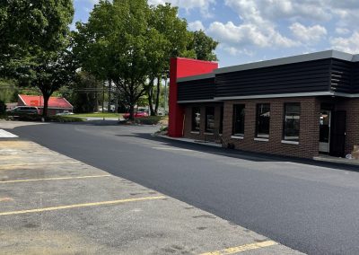 Freshly paved parking lot beside a brick building under a blue sky with scattered clouds and green trees.