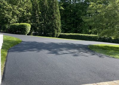 Freshly paved asphalt driveway with green grass, trees, and shrubs under a clear blue sky.