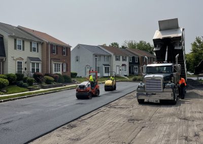 Workers repaving a suburban street with machinery; dump truck and houses line the road.