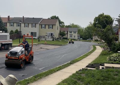 Workers repaving a suburban street with a steamroller; houses and green lawns line the road.